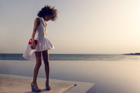 A woman in a white mini-dress and platform shoes standing on a concrete building, standing at an angle and looking down and away from the camera, on the left side of the frame, with water and a peninsula in the background, in the early evening sun.