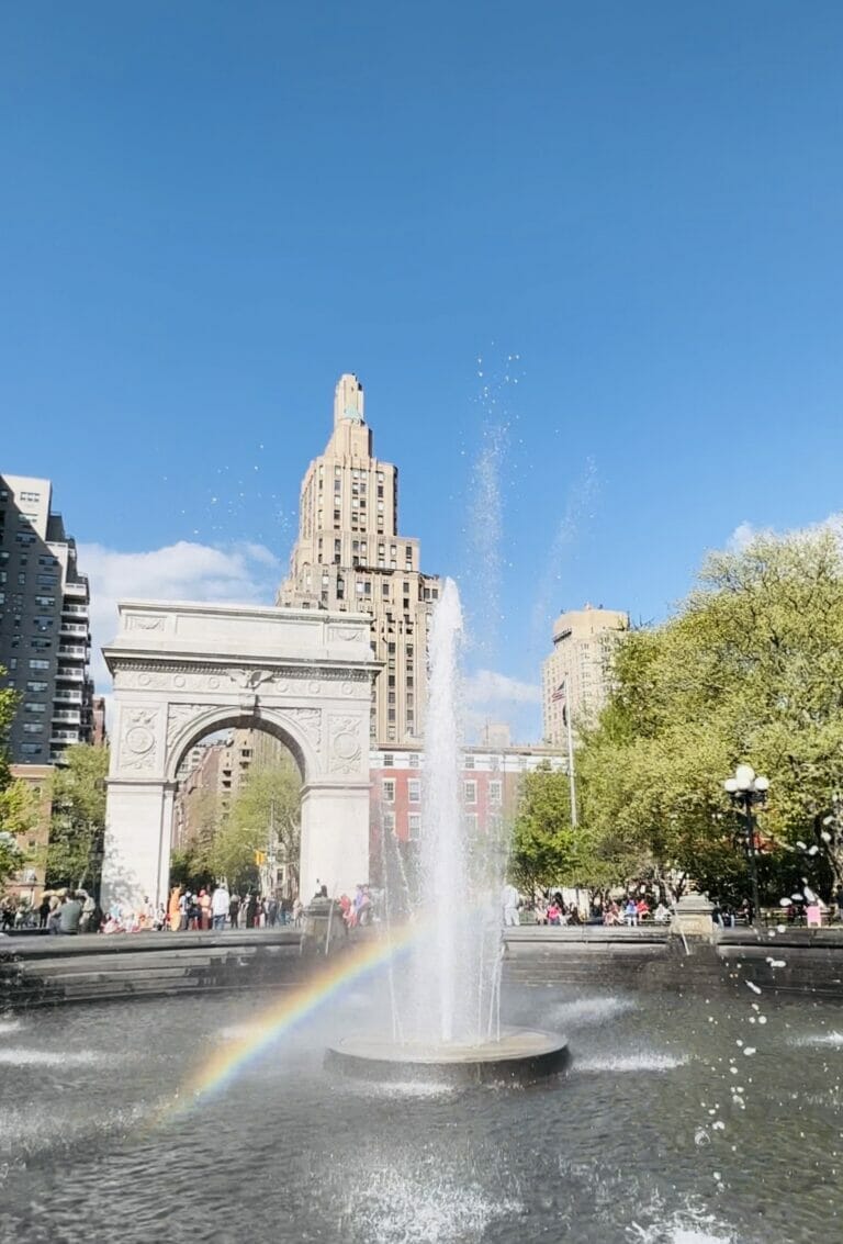 The Washington Square Park fountain on a sunny day creating a rainbow, with the arch in the background