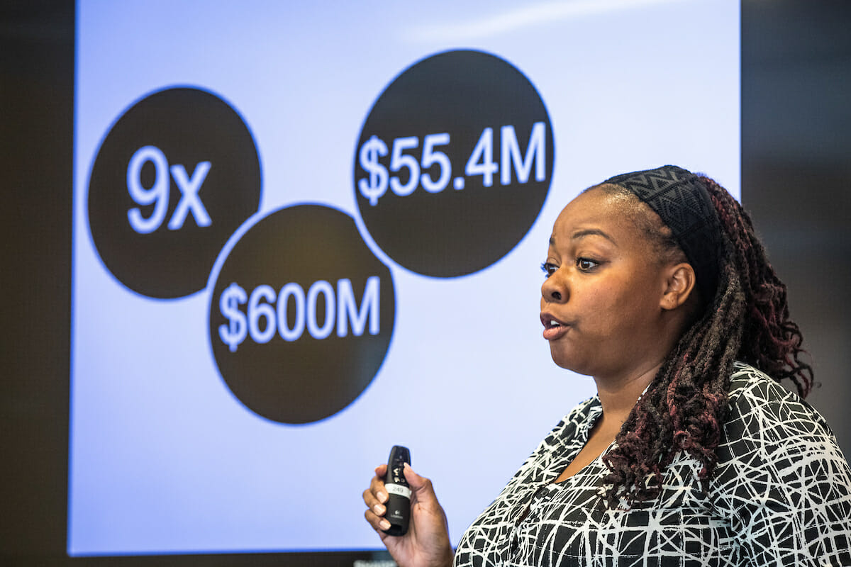 Woman standing in front of a presentation screen, the screen shows numbers and dollar amounts
