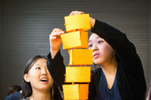 Two high school girls look up at a tower they are building out of yellow cards and clips